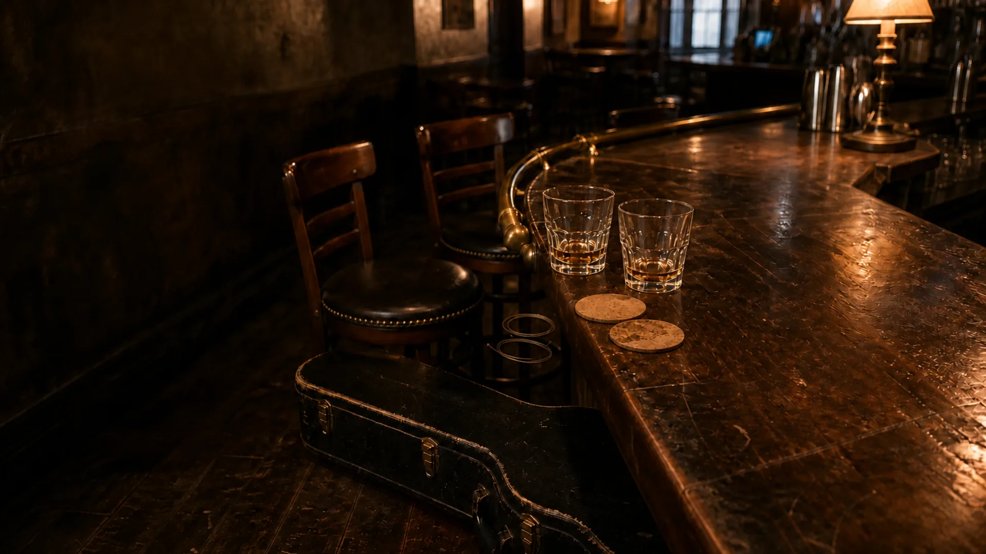 A worn wooden saloon bar holds two glasses and cork coasters, with brass details, pulled-back stools, and a guitar case in a warmly lit room.
